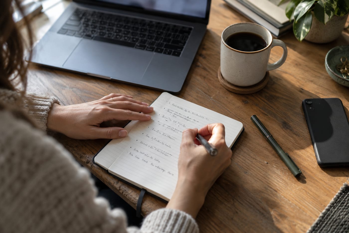 Hands writing structured bullet-point notes in a ruled notebook, coffee mug alongside, laptop blurred in background.