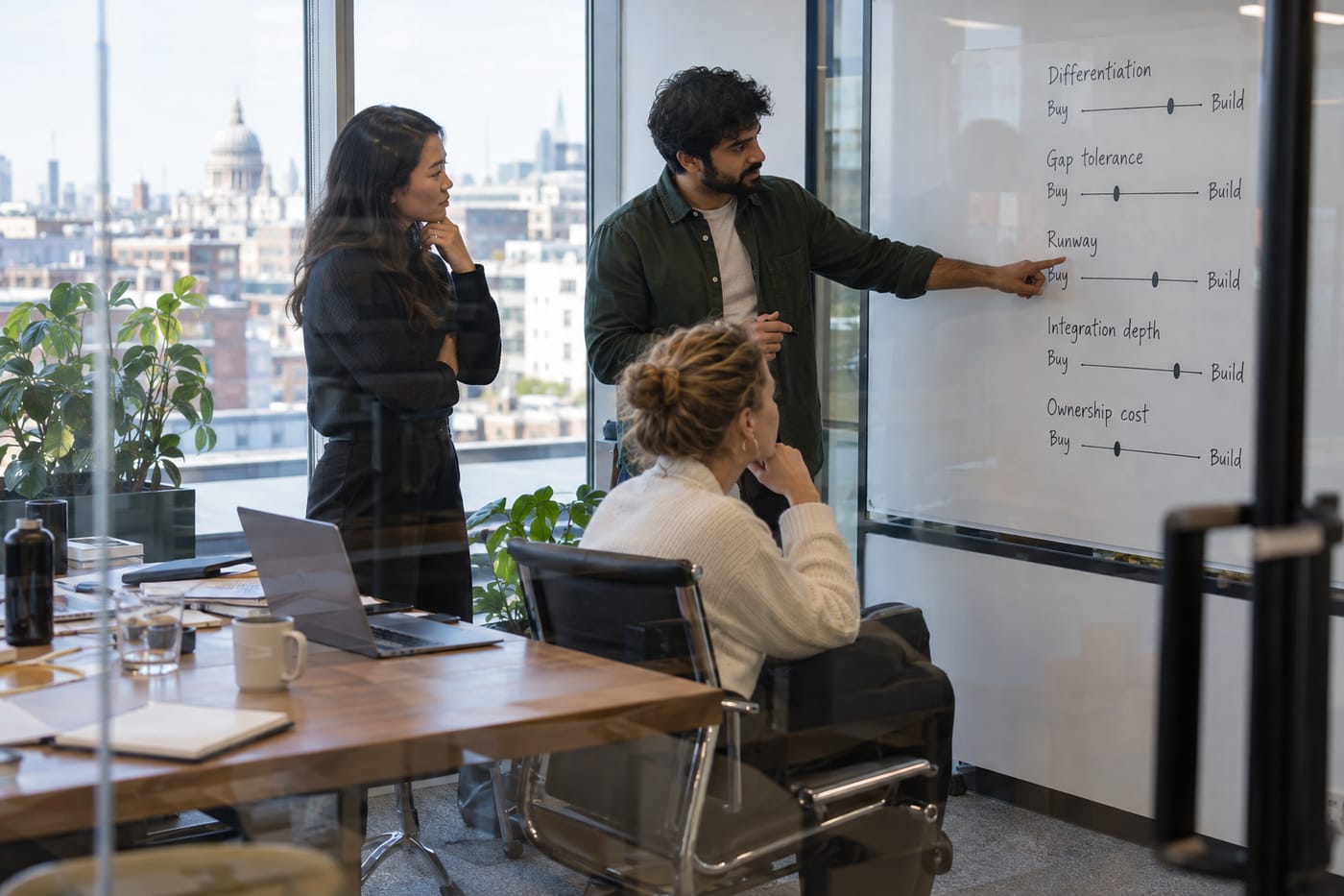 Three UK startup team members scoring five build-vs-buy decision axes on a whiteboard in a glass-walled meeting room.